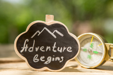 Adventure begins written on a small sign with compass outdoors. Start your journey concept. Green blurred backgroundの写真素材