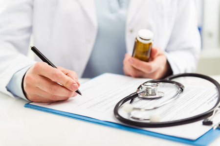 Female doctor filling medical form on clipboard holding ballpoint and medicine bottle. Healthcare and insurance concept.