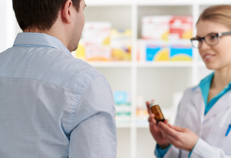Male customer buying pills in drug store. Young female pharmacist consulting client, holding medicine bottle.の写真素材