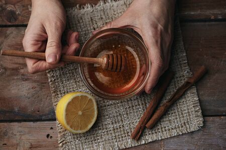 Close-up of female hands holding honey spoon on wooden table with lemon and cinnamon. Natural cold treatment concept.の写真素材