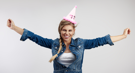 Conceptual shot of smiling birthday female in pink cap with outstretched arms posing in studio.の写真素材