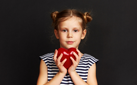Adorable girl holding red heart over dark background. Concept of love, help and protection.の写真素材