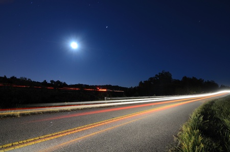 Cars pass on a country road at night with the moon above