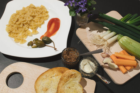 Kitchen table with plate, wooden boards, vegetables, slice of bread, pasta bow and violets. Healthy food concept.の写真素材