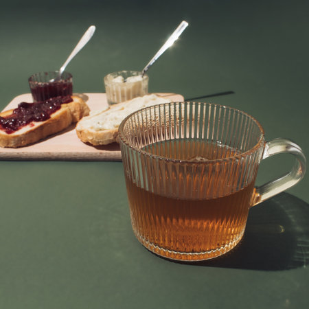 A cup of green tea. Slices of bread on a wooden board spread with cherry jam and butter behinde. Breakfast concept.の写真素材