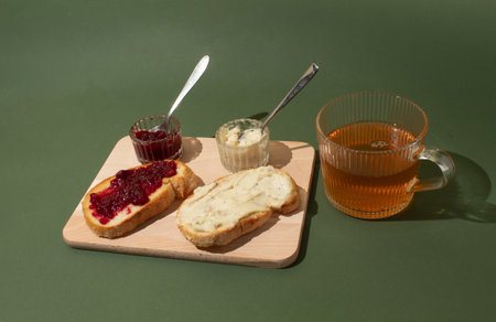 Slices of bread on a wooden board spread with cherry jam and butter. A cup of green tea. Breakfast concept.の写真素材