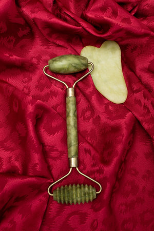 Stone and roller for facial massage gua sha green jade on silk red background, close upの写真素材