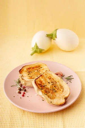 Roasted white eggplant on a pink plate on a pastel yellow background with spices. Creative composition of contemporary foodの写真素材