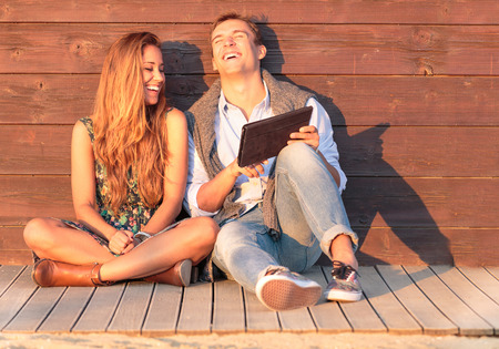 Cheerful guy with girl laugh about video on the tablet. Best friends having fun at the beach with social media and funny contents. Friendship of young people during holiday life moment at sunset.の写真素材