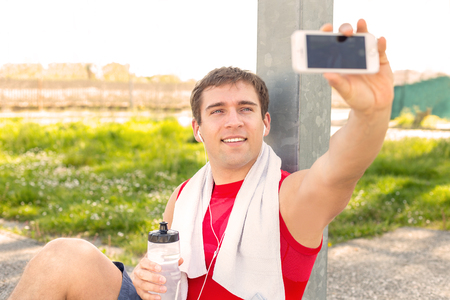 Sitting guy in park and take a selfie during a break at run training with friends - listen music with headphones - Young man finish to do sport and make himself picture - fitness jogging workoutの写真素材