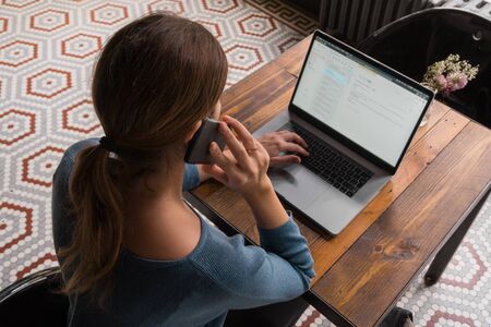 Young freelancer adult sitting on a desk with computer and working. Businesswoman writing email on her notebook and answering the phone. Busy student communicating using laptop during home office.の写真素材