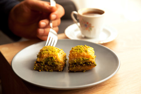 He eats Turkish baklava with green pistachio in a gray plate on an oak coffee table in a home environment, with a fork and Turkish coffee next to itの写真素材