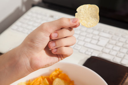 Potato chips in white porcelain plate, caucasian man holding potato chips in hand, snack while working on laptopの写真素材