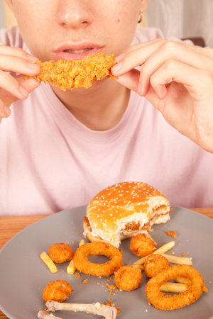 Caucasian man eating golden fried chicken wings, hamburger, potato chips, onion rings and chicken wing pieces in gray plate on wooden table, American fast food cultureの写真素材