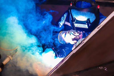 Welder in protective clothing welding metal on a construction site with blue smoke