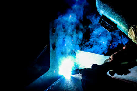 Welder in protective clothing welds metal with electric arc welding machine.