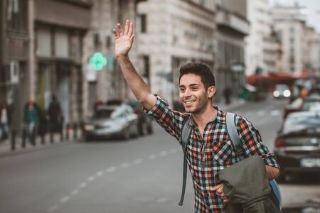 Young man on the street of big city , tries to catch taxi. Carry backpack on one shoulder, street,...