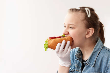 A young girl with blond hair in a blue denim jacket and medical gloves is holding a hot dog and biting it.の写真素材