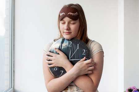 A girl in pajamas and a sleep mask with her eyes closed hugs a large black watch with white hands...