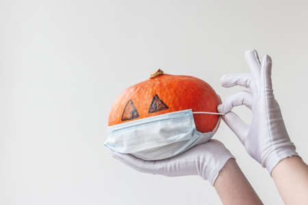 Two white-gloved hands hold an orange pumpkin with painted eyes and put a medical mask on it, safe Halloweenの写真素材