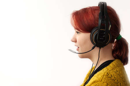A teenage girl with bright red hair, wearing large headphones with a microphone, with her mouth open, she speaks, in a yellow knitted jacket, in profile, looking forward. White background.の写真素材