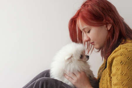 A teenage girl with bright red hair in a yellow, knitted jacket and gray trousers, in profile, holds a white fluffy Pomeranian puppy in front of her face.の写真素材