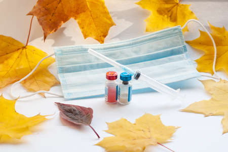 Yellow maple leaves, a medical mask and a syringe in the middle, and two vials of vaccine. The concept of increasing the incidence of diseases in the fall and the need for vaccinationsの写真素材