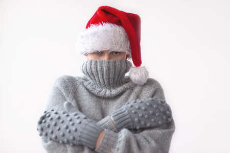 A teenage girl in a gray knitted sweater, mittens and a red Santa hat pulled the collar of the sweater over her face and hugged herself. She was cold. The concept of Christmas, New year and winter.の写真素材