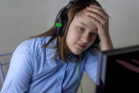 A teenage girl in a blue shirt and headphones looks at the computer monitor, holding her forehead with her hand. Concept of online learning, distance learning, distance work, office work, school.の写真素材