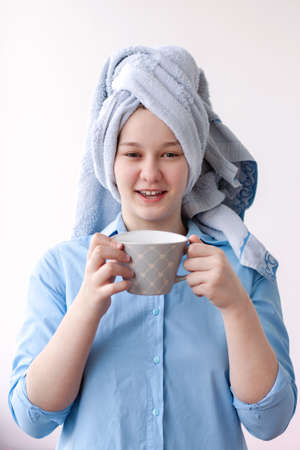 A girl in a blue shirt with a towel on her head holds a large gray cup in her hands. Concept morning, breakfast, tea, coffee, shower.の写真素材
