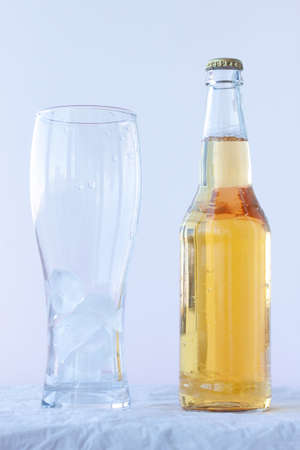 An empty beer glass with ice stands on a white background next to a full bottle of beerの写真素材