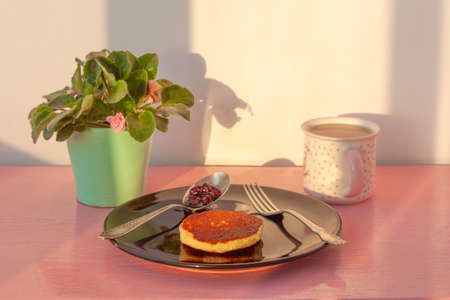 On the pink surface there is a black plate with a pancake, next to it is a fork and a spoon with...