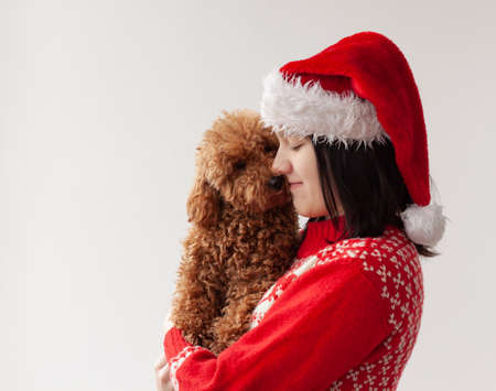 A teenage girl in a Santa Claus hat and a red Christmas sweater holds a miniature poodle in her arms. The concept of Christmas and New Year.の写真素材