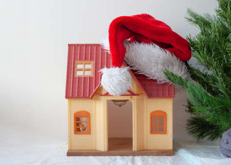 The toy house is wearing a Santa Claus hat next to an artificial Christmas treeの写真素材