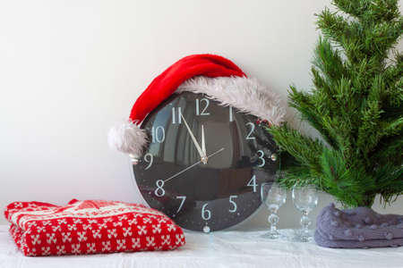A clock in a Santa Claus hat next to an artificial Christmas tree and a Christmas sweaterの写真素材