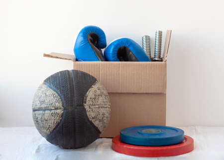 An old basketball and barbells in red and blue next to a cardboard box with boxing gloves