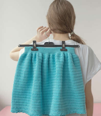 The girl stands with her back and holds a hanger with a knitted blue skirt in her hand.の写真素材