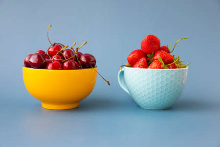 On a blue background there are two bowls with cherries and strawberries