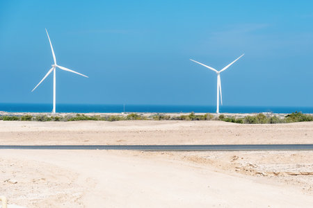 Two wind turbines stand tall against a backdrop of the vast blue ocean and desert landscape. The wind farm captures the essence of renewable energyの写真素材