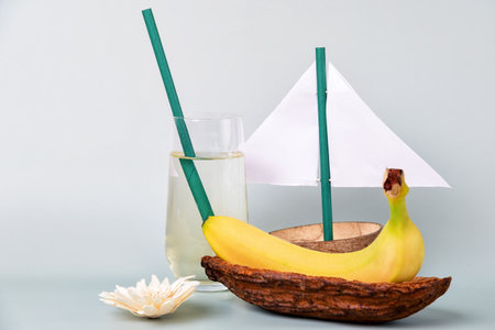 Banana nestled in a cocoa bean pod, creatively arranged to resemble a boat with a paper sail and a straw mast. Glass of water with a straw and flowerの写真素材