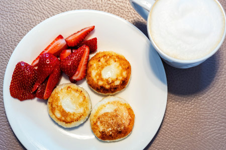 A delicious and healthy breakfast of syrniki served with fresh strawberries and a cup of frothy cappuccino. Traditional Eastern European dishの写真素材