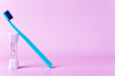 Blue toothbrush rests against a small hourglass timer on a pastel pink background, emphasizing the importance of timed dental hygiene routinesの写真素材