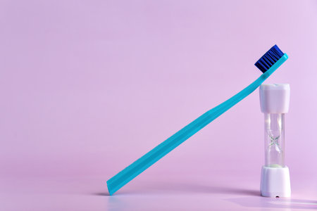 Blue toothbrush leans against a white sand timer on a pastel pink background, illustrating the recommended two-minute oral brushing timeの写真素材