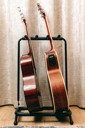 Two acoustic guitars stand side-by-side on a black metal guitar stand in front of beige curtainsの写真素材