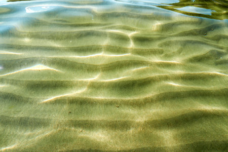 Sunlight dances through clear water, creating mesmerizing patterns on the sandy bottom below. Subtle ripples and textures offer serene visualの写真素材