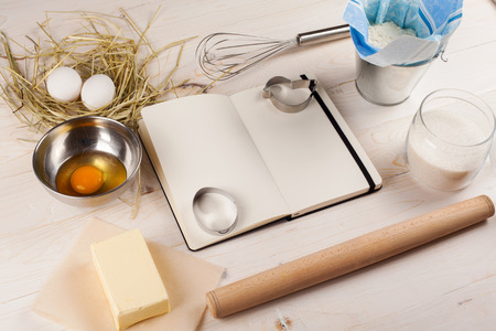 Ingredients for the dough with a book for recipes for Easter. Eggs, flour, butter, sugar and kitchen tools on a white wooden background. Rustic background with free text space.の写真素材