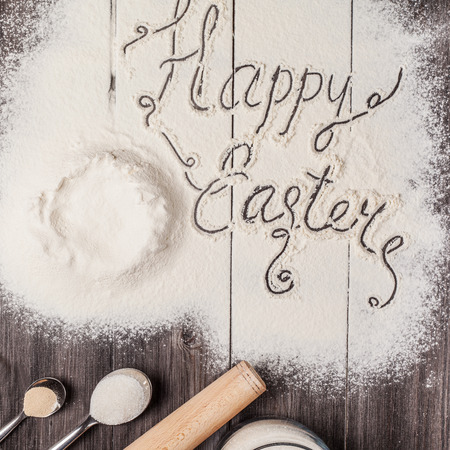 Ingredients for the dough and baking with the words Happy Easter in the center of composition. Eggs, flour, butter, sugar and kitchen tools on a dark wooden background. Rustic background. Flat lay.の写真素材