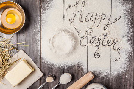 Ingredients for the dough and baking with the words Happy Easter in the center of composition. Eggs, flour, butter, sugar and kitchen tools on a dark wooden background. Rustic background. Flat lay.の写真素材