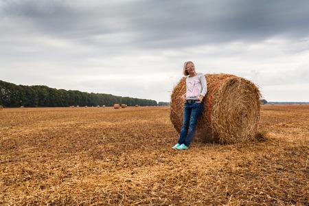 A young woman stands on the field, leaning on a sheaf of hay.の写真素材