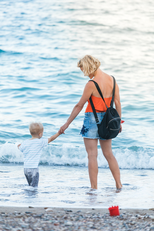 A young and beautiful mother walks in the fresh air on the beach with her young child. The boy squatted down to get a pebble he liked at sunset time. The concept of a happy family, lifestyle, vacation at sea, walking in the fresh air with childrenの写真素材